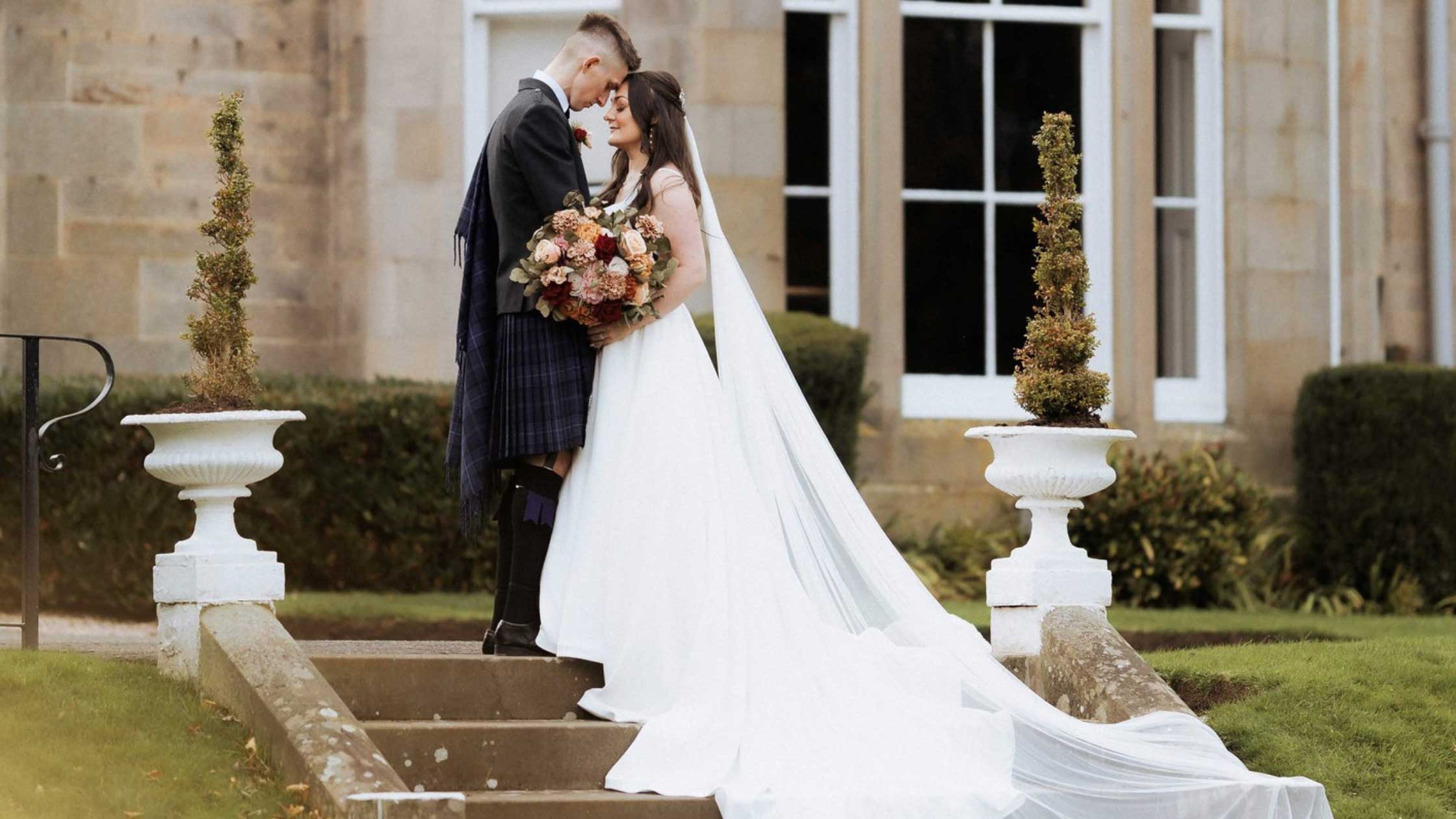 A bride in a long white wedding gown with a flowing train and veil stands on stone steps outdoors, kissing a groom in a dark formal suit. The setting includes manicured hedges, white planter boxes, and a stone building with tall windows in the background.